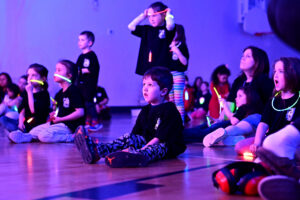 Students watching the concert, all a-glow with glowstick necklaces and bracelets in a black lit gym.
