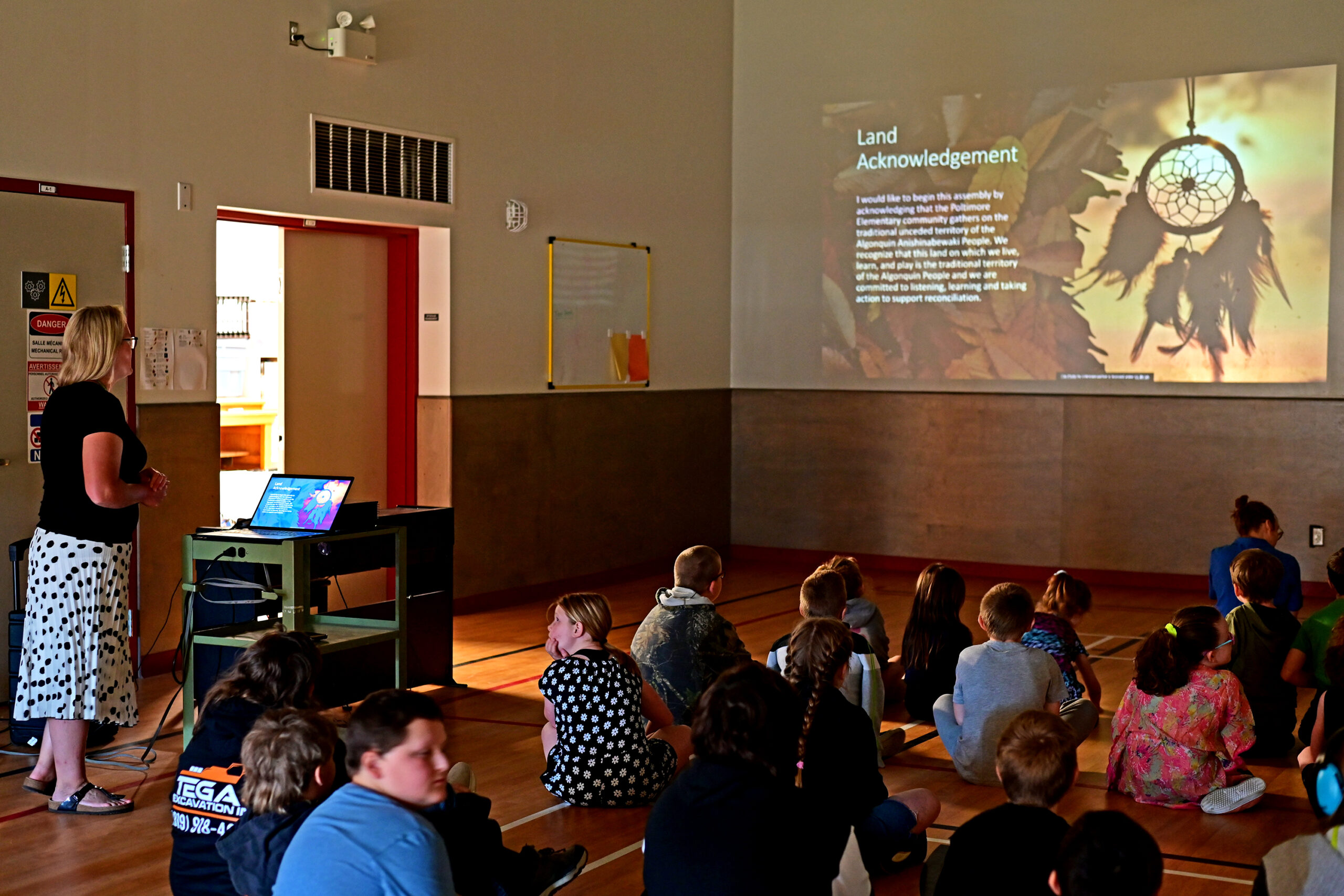 Poltimore principal Melanie Hogan standing at a podium with a land acknowledgement projected on the gymnasium wall